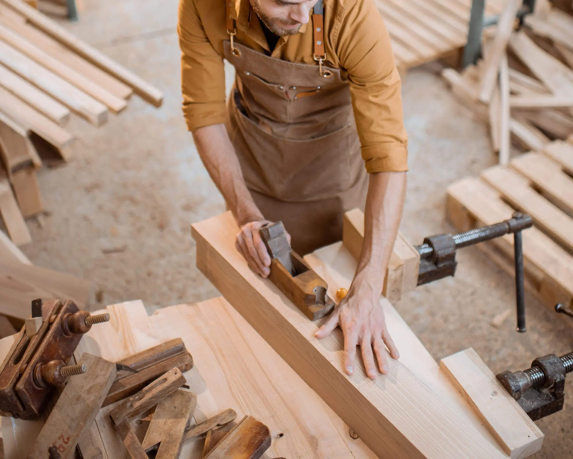 Skilled Carpenter Shaping Wood in Workshop A carpenter using a hand plane to smooth a piece of wood in a woodworking workshop, surrounded by various wooden planks and tools.