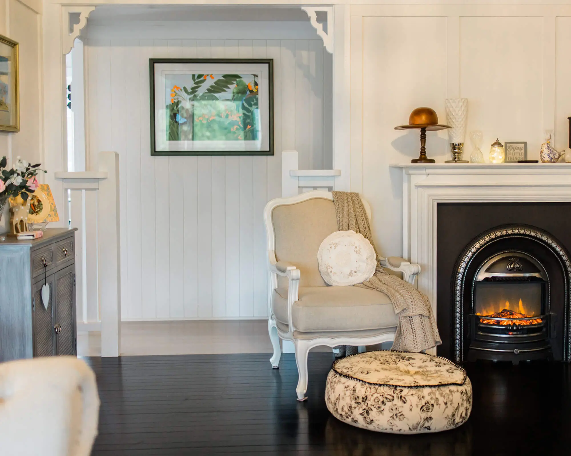 Cozy Living Room with Fireplace and Stylish Decor A beautifully styled living room featuring a comfortable chair, a decorative pouf, and a glowing fireplace, complemented by artwork on the wall.