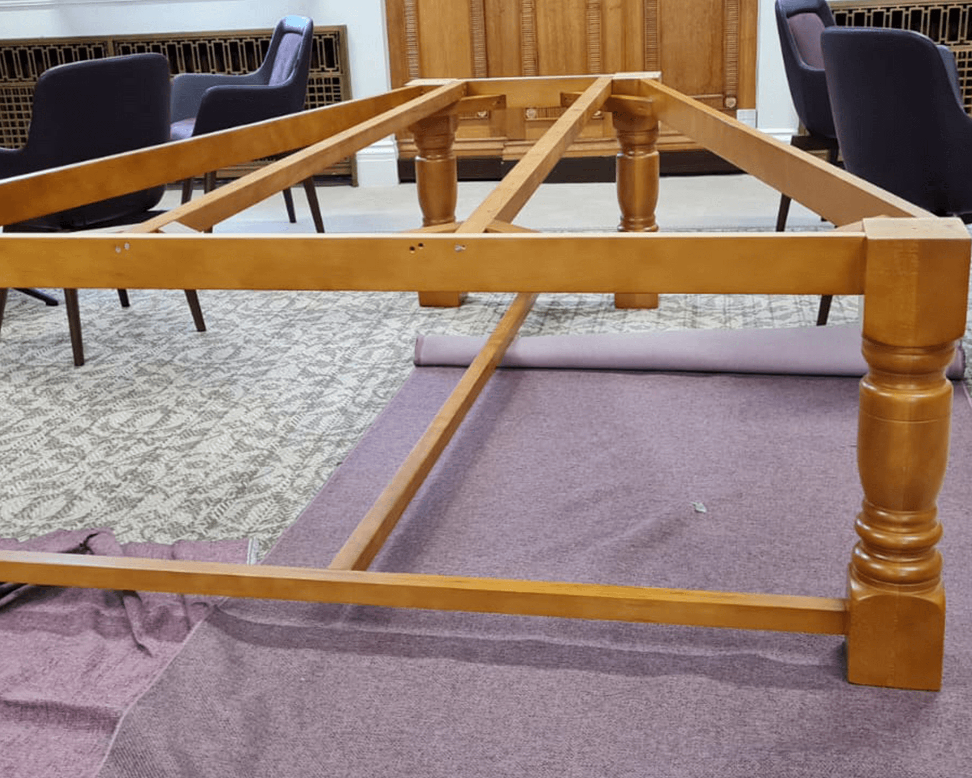 An unfinished wooden table frame displayed upside down on a carpeted surface, showcasing the structural design and craftsmanship.