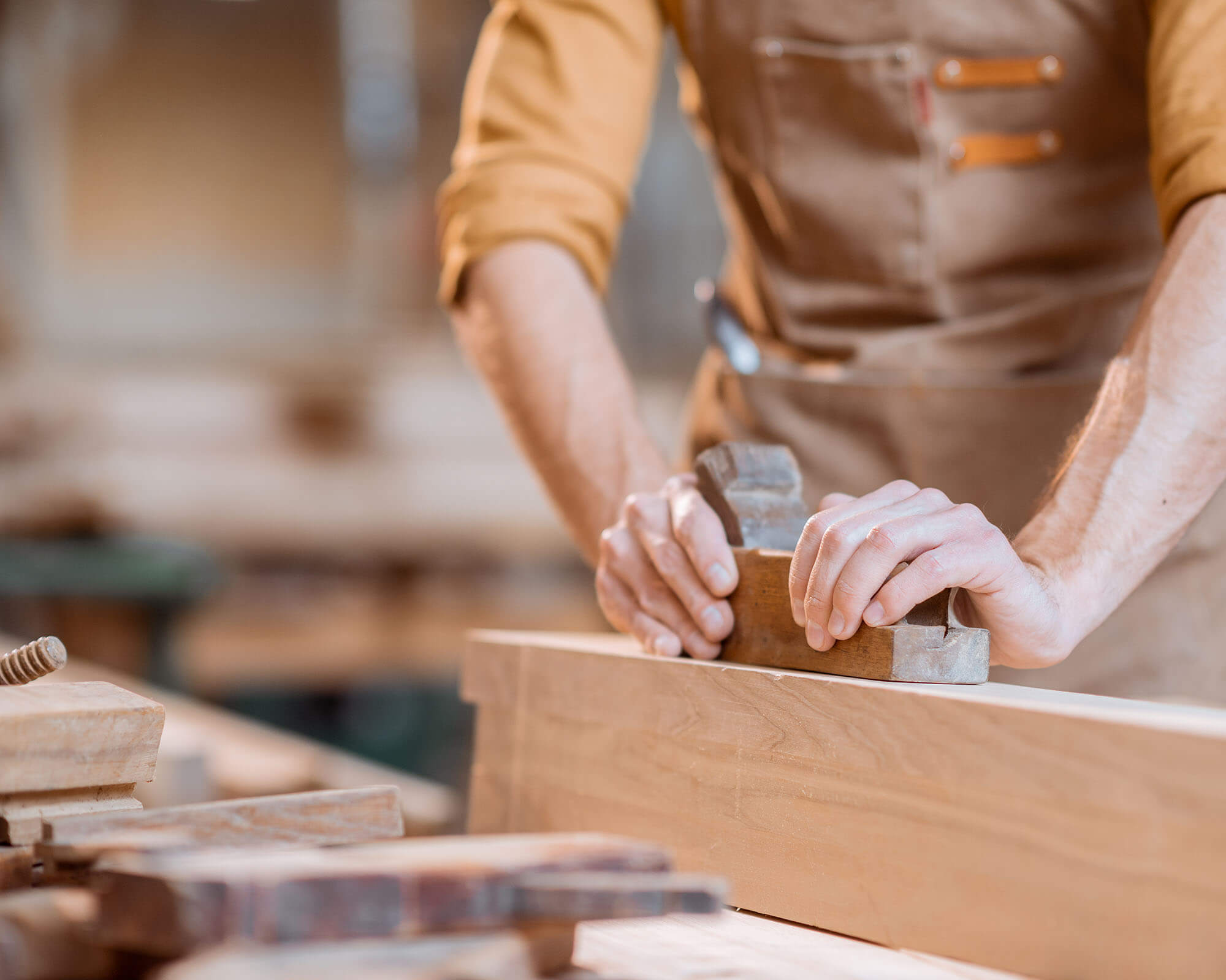 Close-up of a craftsman's hands using a hand plane to smooth a wooden plank in a workshop setting.