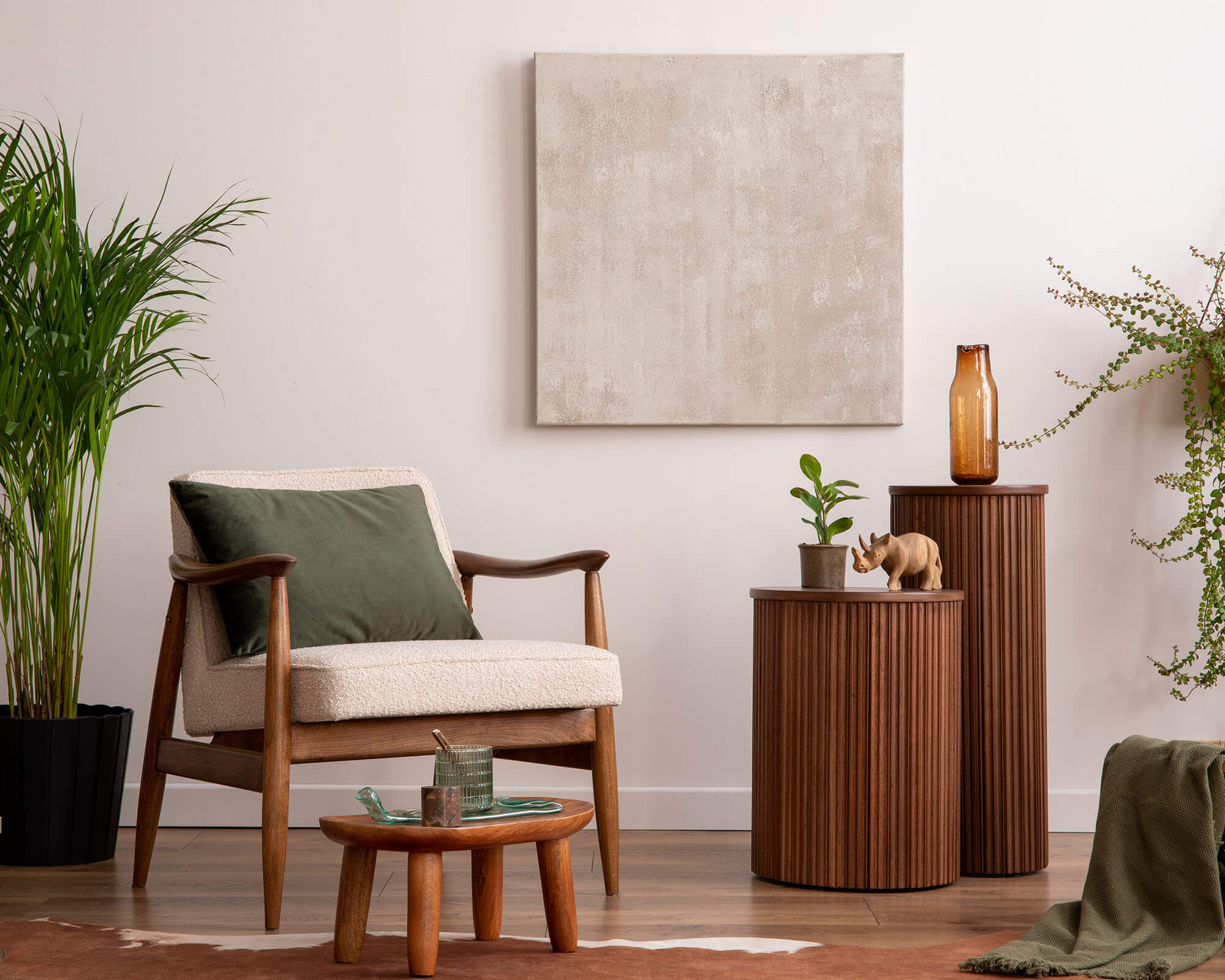 A cozy living room featuring a wooden armchair, decorative tables, and indoor plants, complemented by a neutral artwork on the wall.