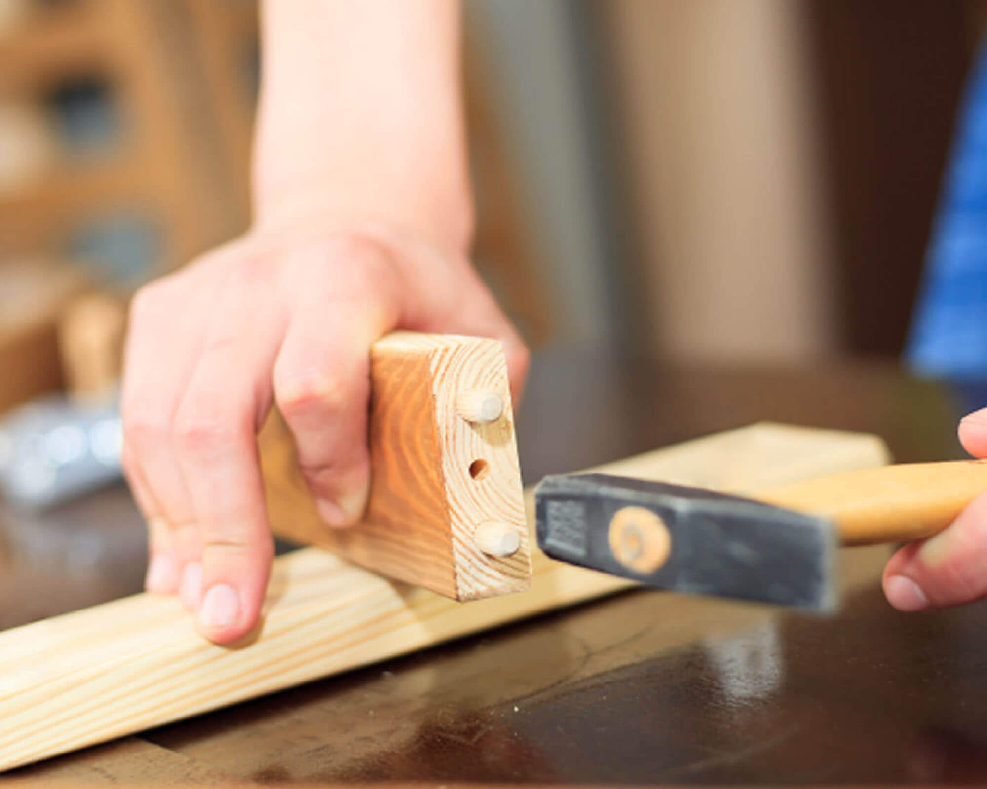 A close-up of a hand holding a wooden piece while using a hammer to assemble it on a workbench.